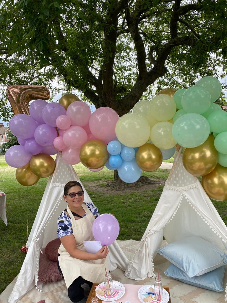Ela kneeling in front of two teepees, with support golden and pastel coloured balloons bridging across them