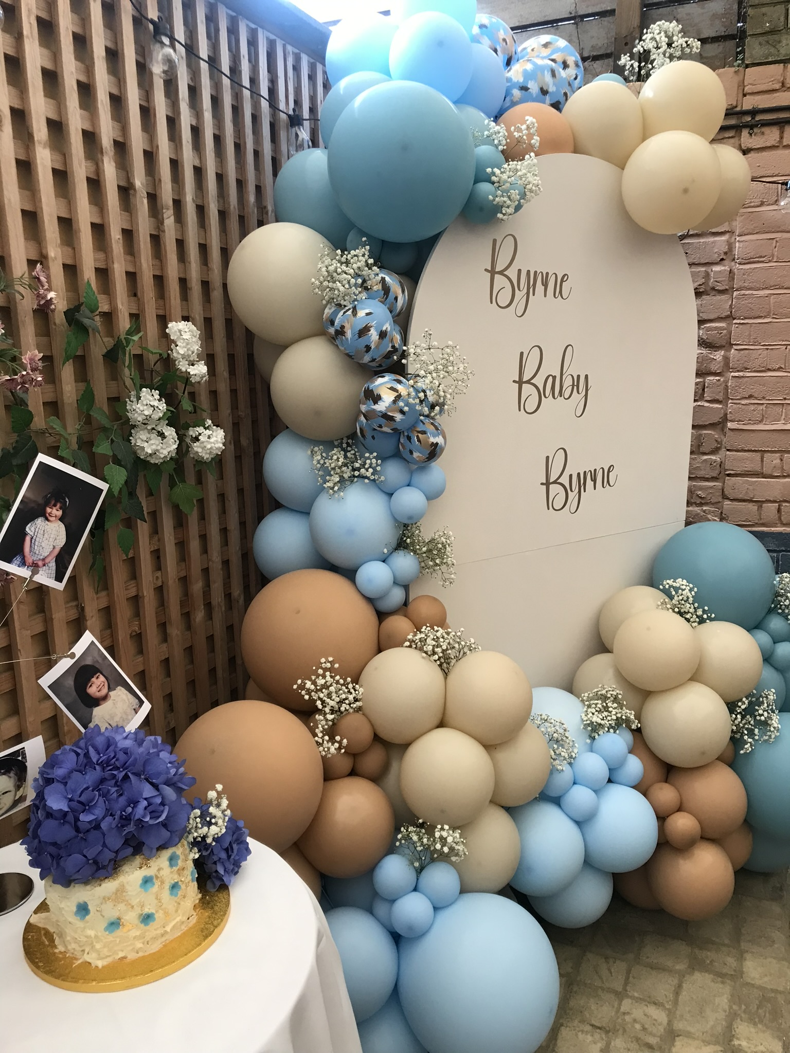 A sailboard with a cursive golden decal saying 'Byrne Baby Byrne' is surrounded by blue and beige balloons