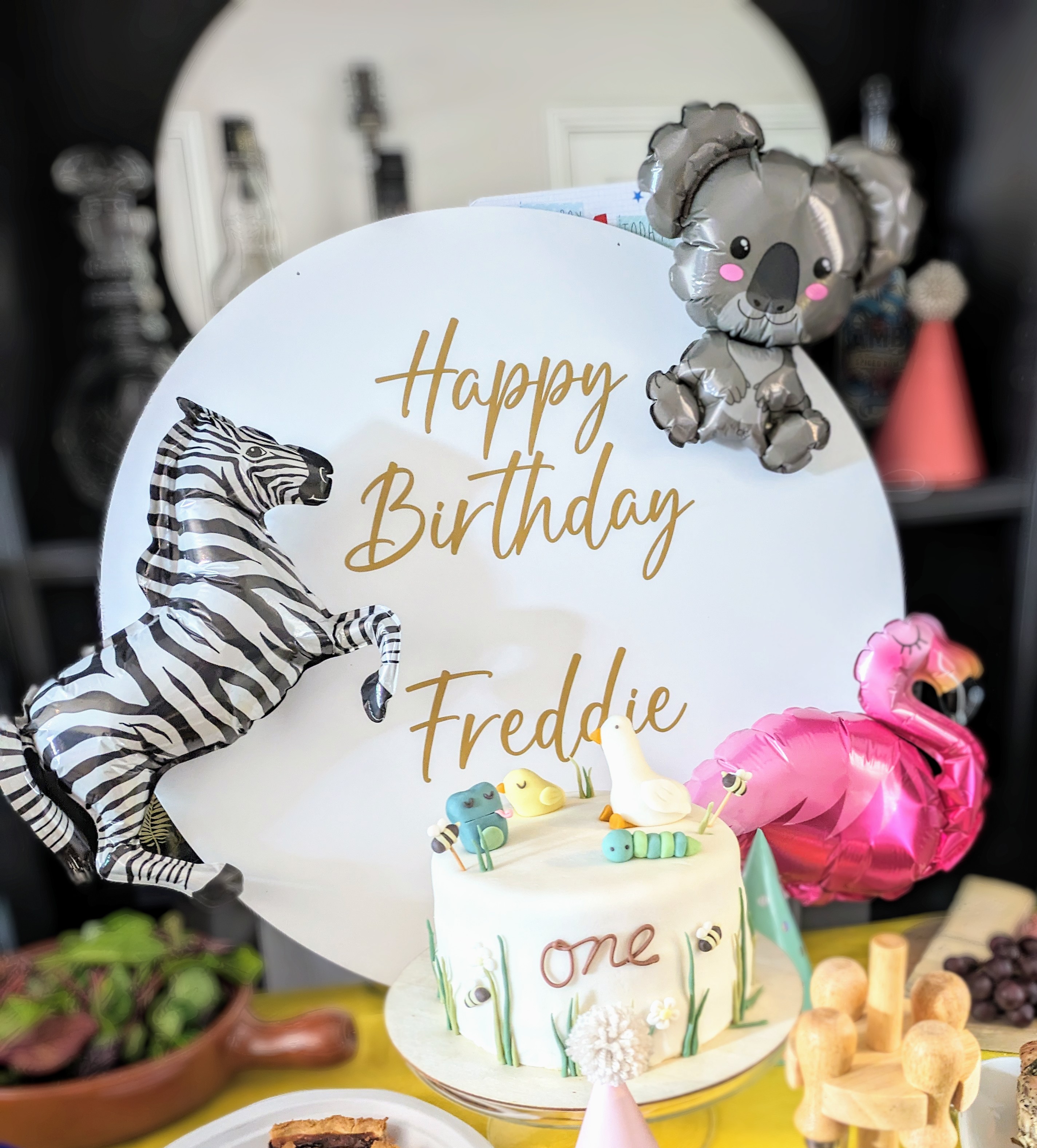 A white acrylic disc with a Happy Birthday message written in cursive gold letters sits behind a cake, surrounded by foil balloons of exotic animals