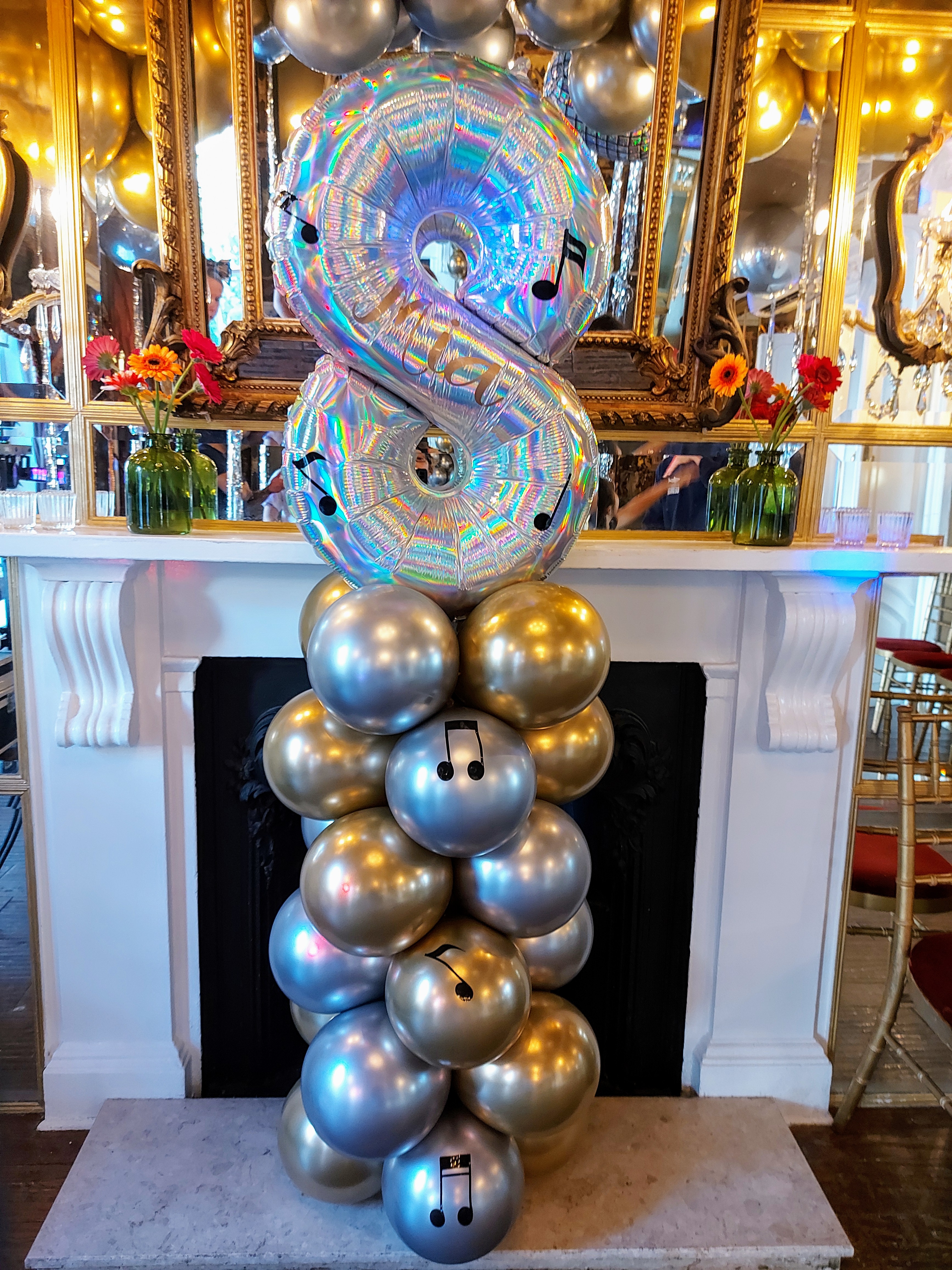 A silver and gold balloon column decorated with musical notes supports a large iridescent number 8