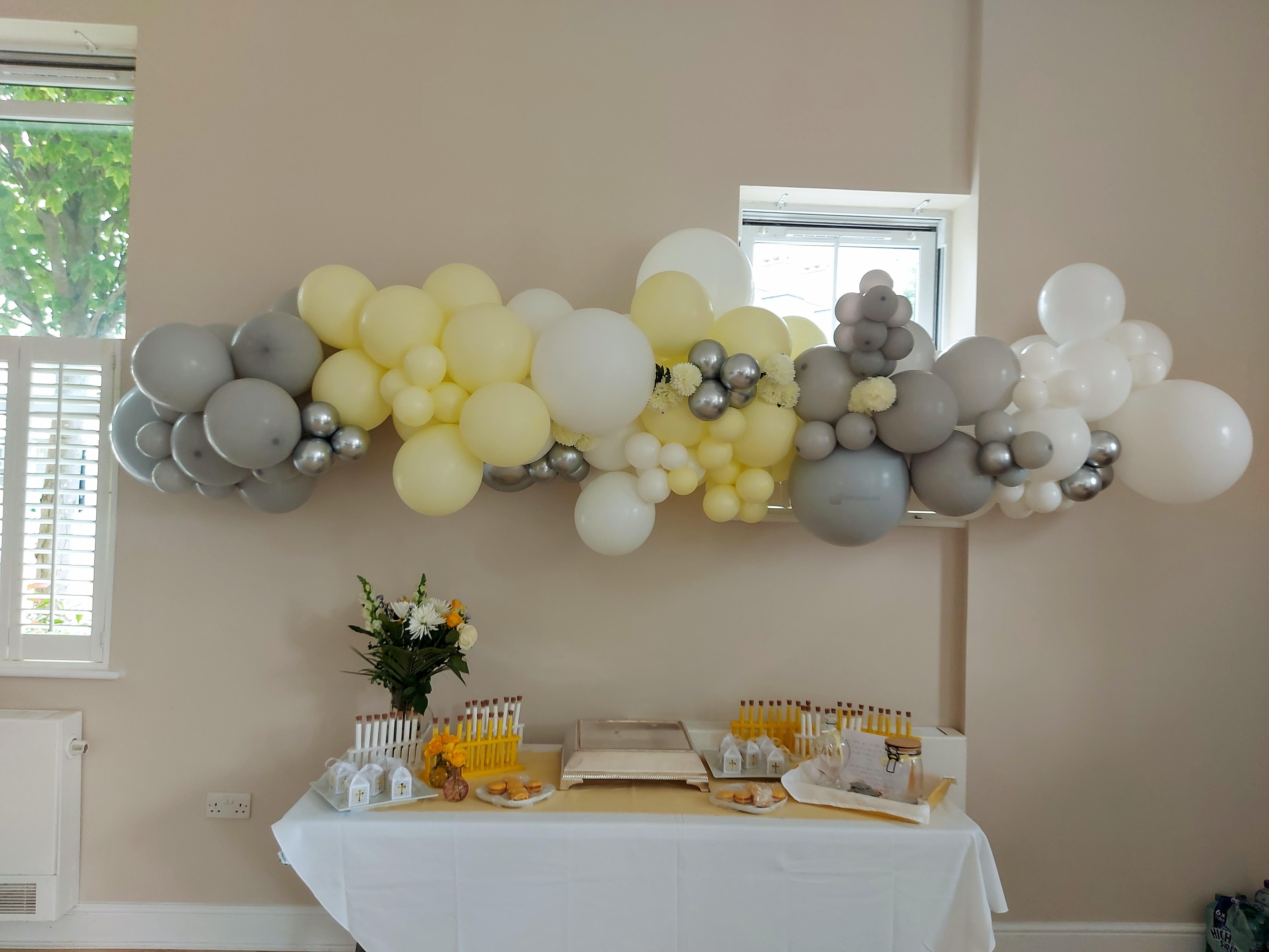 An organic garland of white, yellow and silver balloons adorns a baptism shrine