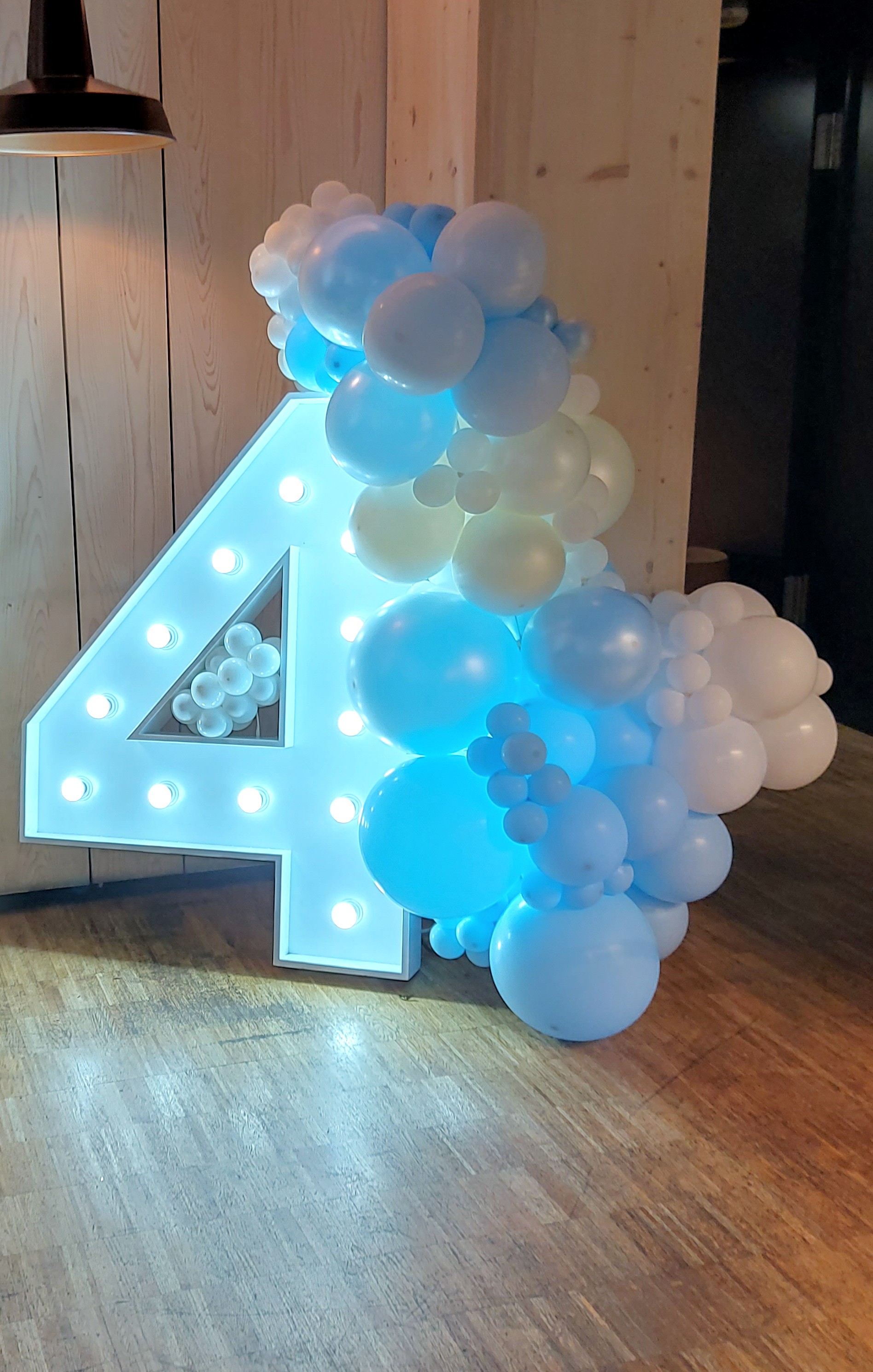 A large white wooden number 4 lit up by LEDs is supported by a wave of organic blue and white balloons