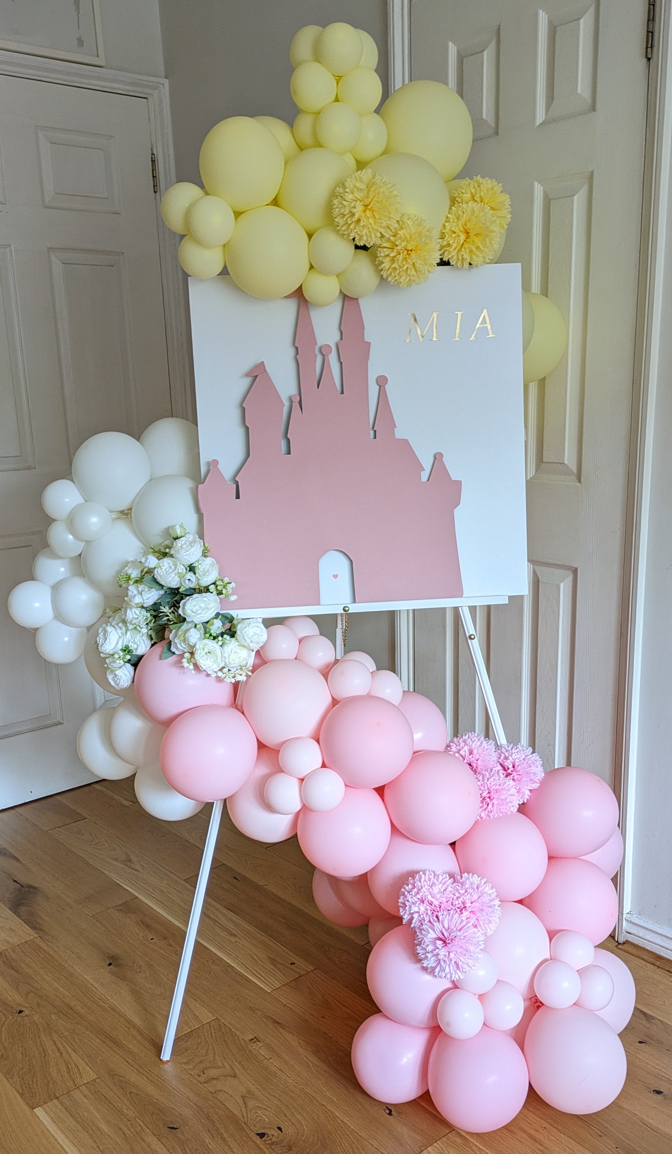A flowery balloon stack coils around an easel holding up a pink cutout of a fairy castle